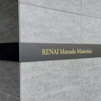 壁面についている、L字型プレート型館銘板。曲げは左側。一面黒いプレートの中央に、光るタイプの物件名の文字がある。