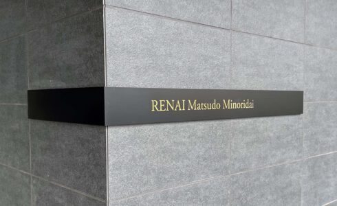 壁面についている、L字型プレート型館銘板。曲げは左側。一面黒いプレートの中央に、光るタイプの物件名の文字がある。