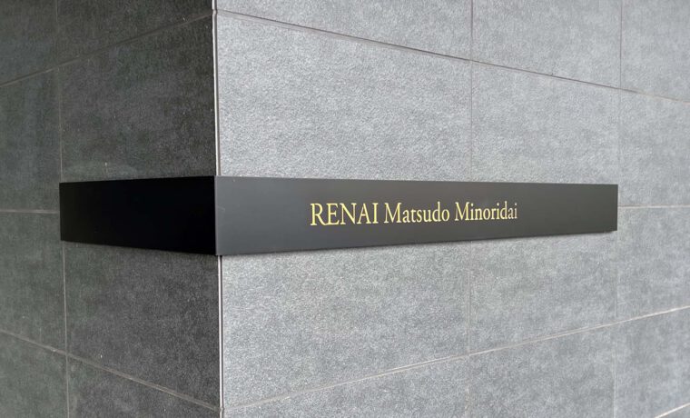 壁面についている、L字型プレート型館銘板。曲げは左側。一面黒いプレートの中央に、光るタイプの物件名の文字がある。