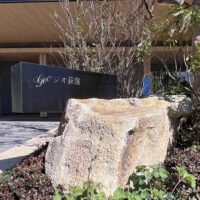 マンションアプローチ館銘板。 白い大きな岩の上部に、やや食い込むような形で横に長い長方形の黒い館銘板の箱が乗っている。箱の左上には、内側から光るタイプの物件名の文字がある。