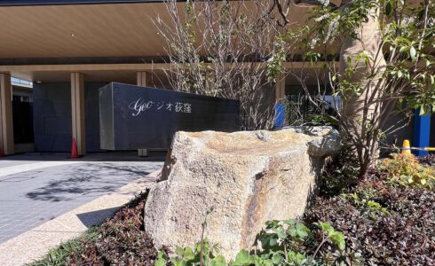 マンションアプローチ館銘板。 白い大きな岩の上部に、やや食い込むような形で横に長い長方形の黒い館銘板の箱が乗っている。箱の左上には、内側から光るタイプの物件名の文字がある。