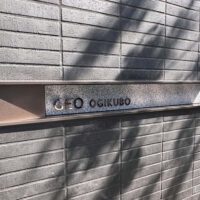壁面についている、L字型プレート型館銘板。曲げは右側。 ブロンズのフレームの内側に、御影石のベースがあり、その上にブロンズの切文字の物件名がある。