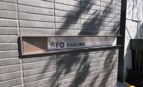 壁面についている、L字型プレート型館銘板。曲げは右側。 ブロンズのフレームの内側に、御影石のベースがあり、その上にブロンズの切文字の物件名がある。