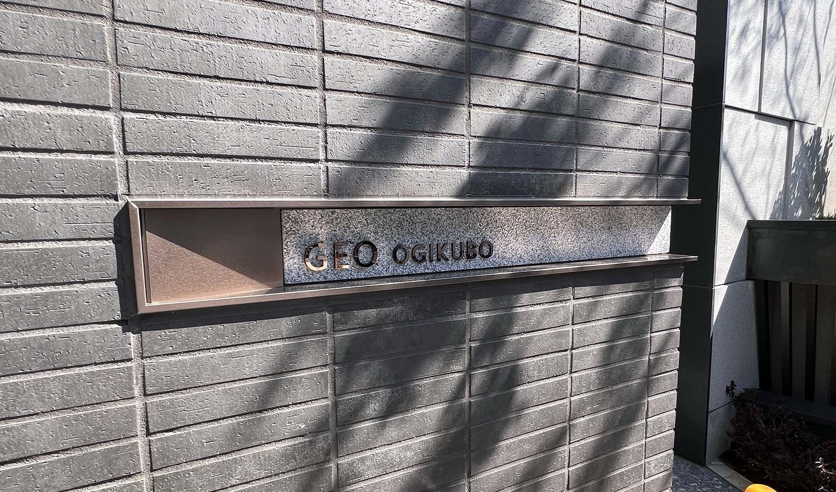 壁面についている、L字型プレート型館銘板。曲げは右側。 ブロンズのフレームの内側に、御影石のベースがあり、その上にブロンズの切文字の物件名がある。