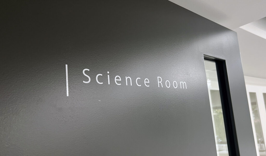 黒い壁についている、白いカッティングシールの切文字室名表示。「Science Room」と書かれており、頭の文字の左側には白い縦のラインが入っている。