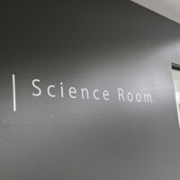 黒い壁についている、白いカッティングシールの切文字室名表示。「Science Room」と書かれており、頭の文字の左側には白い縦のラインが入っている。