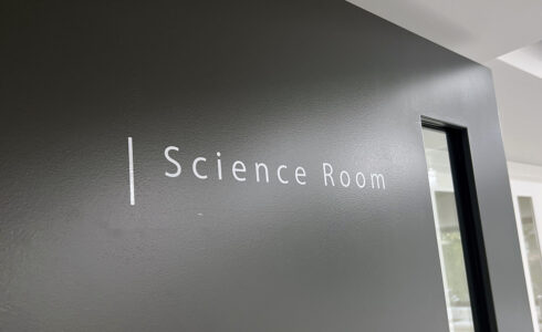 黒い壁についている、白いカッティングシールの切文字室名表示。「Science Room」と書かれており、頭の文字の左側には白い縦のラインが入っている。