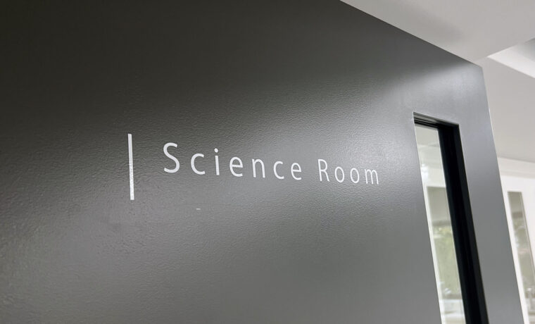 黒い壁についている、白いカッティングシールの切文字室名表示。「Science Room」と書かれており、頭の文字の左側には白い縦のラインが入っている。