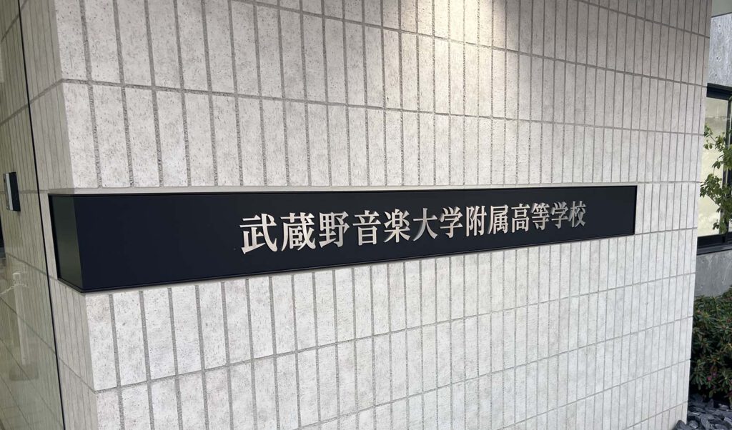 壁面についている、L字型プレート型館銘板。曲げは左側。一面黒いプレートの中央に、ステンレスの切文字の物件名がある。