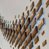 壁面アート照明の拡大写真。 菱形の木製パネルが波状に壁に取り付けられており、その背面から発光パターンをプログラムされたLEDライトが、様々な色で発光している。