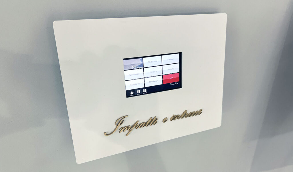 壁面アート照明操作板。 壁についた白いボックスの中央に、操作用のタッチパネルがある。その下には「Impatti e intrecci」と書かれたゴールド色のアクリル切文字が付いている。