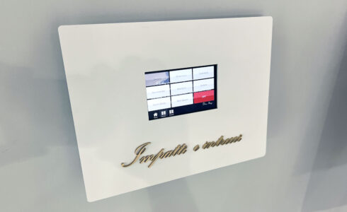 壁面アート照明操作板。 壁についた白いボックスの中央に、操作用のタッチパネルがある。その下には「Impatti e intrecci」と書かれたゴールド色のアクリル切文字が付いている。