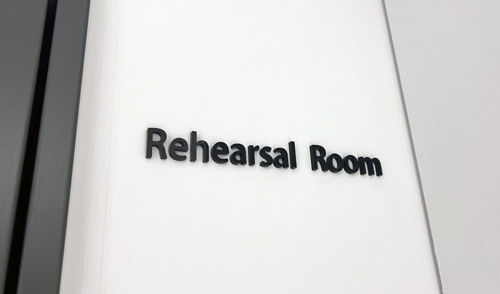 白い壁についている、黒のアクリル切文字室名表示。「Rehearsal Room」と書かれている。
