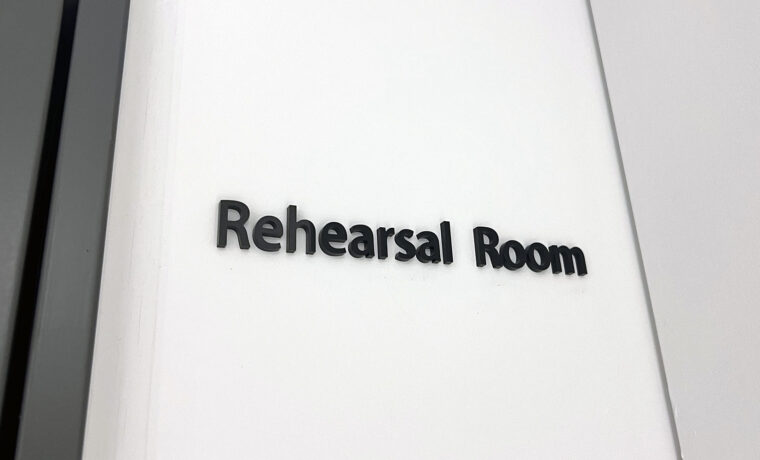 白い壁についている、黒のアクリル切文字室名表示。「Rehearsal Room」と書かれている。