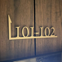 木目調の壁についている、住戸案内表示。ステンレス切文字で色はゴールド。「101-102」と書かれた部屋番号の下に横線が引かれており、線の左端は90度上に曲げられ、先端が矢印になっている。
