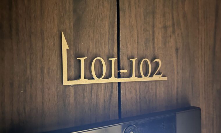 木目調の壁についている、住戸案内表示。ステンレス切文字で色はゴールド。「101-102」と書かれた部屋番号の下に横線が引かれており、線の左端は90度上に曲げられ、先端が矢印になっている。
