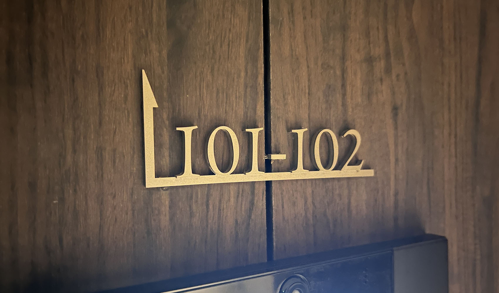 木目調の壁についている、住戸案内表示。ステンレス切文字で色はゴールド。「101-102」と書かれた部屋番号の下に横線が引かれており、線の左端は90度上に曲げられ、先端が矢印になっている。